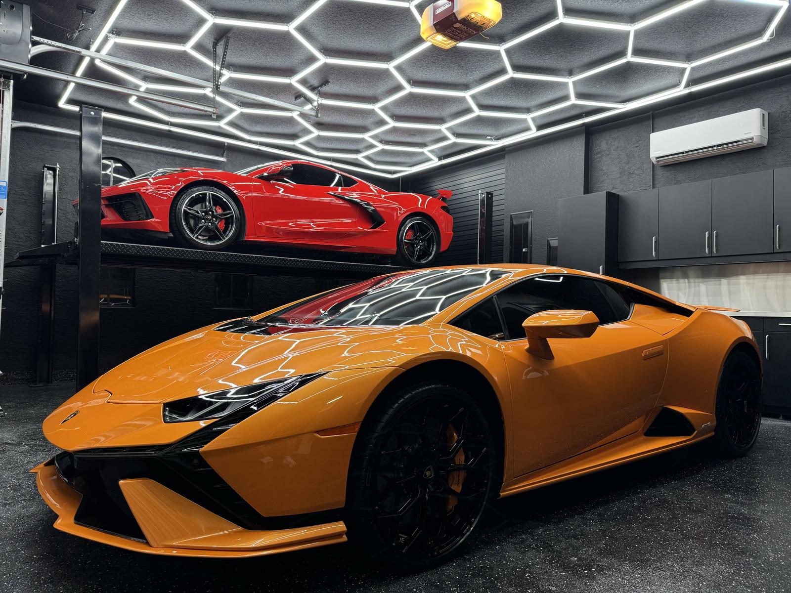 An orange Lamborghini parked under a red Corvette raised on a STAK four-post lift in a home garage
